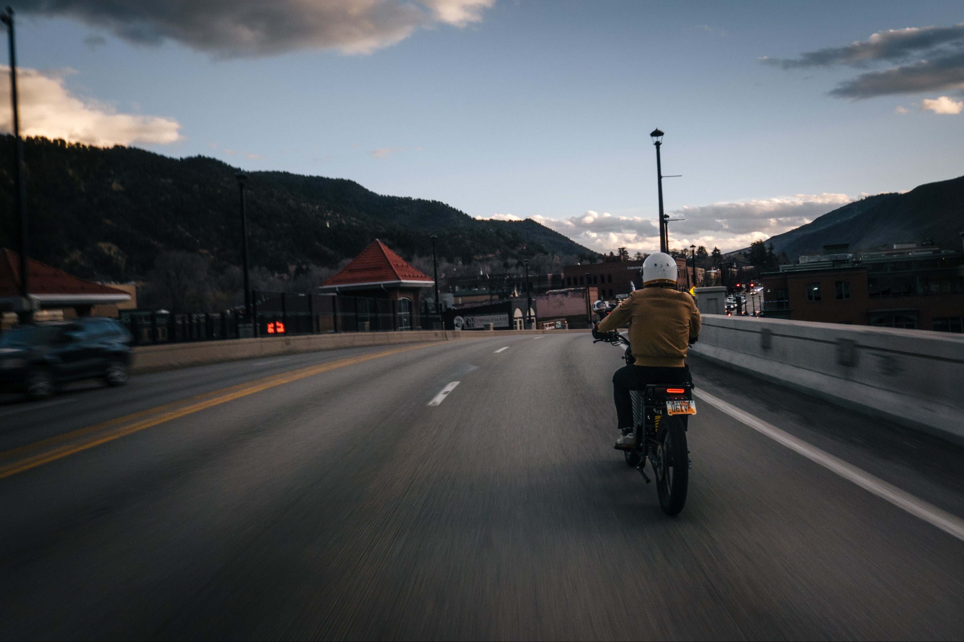 Person riding a motorcycle on a road with a cityscape in the background during sunset.